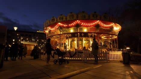 Carrusel-O-Tiovivo,-Girando-En-La-Oscuridad,-Las-Madres-De-Los-Niños-Mirándolos-Y-La-Gente-Caminando,-En-El-Festival-De-Navidad-De-Sofía-2023