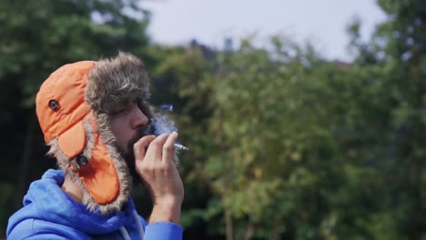 Un-Hombre-Con-Una-Sudadera-Azul-Fuma-Y-Lleva-Un-Cálido-Sombrero-De-Aviador-De-Invierno-De-Color-Naranja