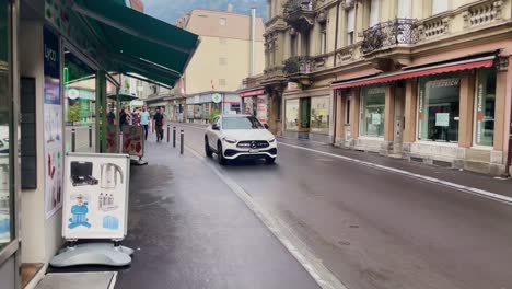 Car-Passing-Eloquent-Buildings-Downtown-|-Interlaken-Switzerland-Immersive-Travel-Tourism-Mountainside-Valley-Resort-City,-Europe,-Walking,-Rainy-Day,-4K