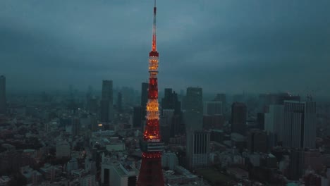 Drohnenantenne-über-Der-Stadt-Tokio,-Die-In-Der-Abenddämmerung-In-Richtung-Des-Von-Hohen-Wolkenkratzern-Umgebenen-Tokio-Towers-Schwenkt,-Mit-Stimmungsvollen-Dunklen-Wolken-Und-Einem-Smogverschmutzten-Himmel