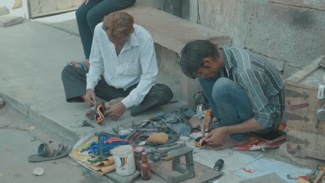 two-indian-men-sitting-on-the-street-in-New-Delhi,-India-tailoring-a-pair-of-shoes