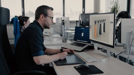 Un-Hombre-Con-Gafas-Sentado-Frente-A-La-Computadora-Trabajando-En-El-Concepto-De-Diseño-De-Un-Producto-Moderno-En-La-Oficina-Rodeado-De-Colegas-En-Una-Joven-Empresa-Emergente