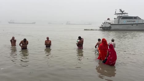 Vista-Estática-De-Hombres-Y-Mujeres-Bañándose-Y-Rezando-En-Una-Mañana-De-Invierno-Durante-El-Festival-Sankranti-Con-Embarcadero-Al-Fondo-En-Babughat,-Kolkata.