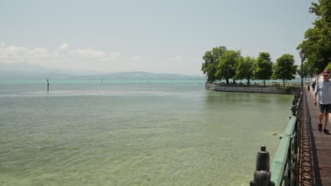 Man-strolling-by-the-calm-waters-of-Meersburg,-Bodensee,-with-mountains-in-the-distance,-clear-sky