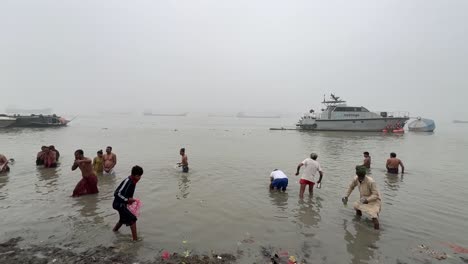 Hombres-Y-Mujeres-Bañándose-Y-Rezando-En-La-Brumosa-Mañana-De-Invierno-Durante-Sankranti-Con-Embarcadero-Al-Fondo-Durante-Una-Tarde-Fría-En-Babughat,-Kolkata