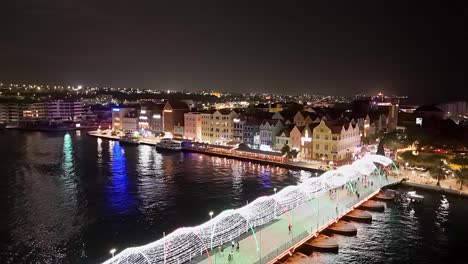 Handelskade-Willemstad-Luces-Brillantes-Se-Reflejan-En-El-Agua-Sobre-El-Puente-Reina-Emma,-Descenso-Aéreo