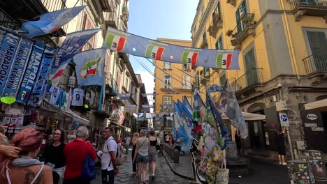 Toma-En-Primera-Persona-De-Caminar-Por-Una-Calle-Estrecha-Y-Colorida-Con-Un-Símbolo-Del-Fútbol-En-El-Centro-De-La-Ciudad-De-Nápoles,-En-Italia,-Llena-De-Turistas.