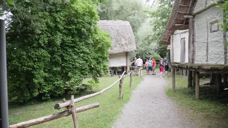 Grupo-De-Personas-Recorriendo-Cabañas-Tradicionales-Con-Techo-De-Paja-En-Un-Museo-De-La-Edad-De-Hielo,-Rodeado-De-Exuberante-Vegetación