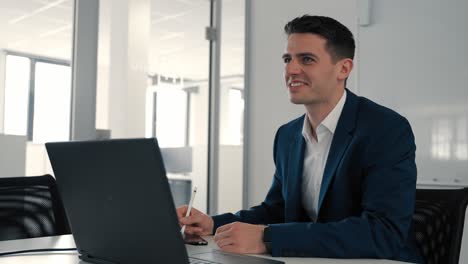 Young-smiling-caucasian-businessman-sitting-at-a-desk-in-front-of-a-camputer-having-a-video-call-and-making-notes-in-an-office-room