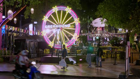 View-from-the-back-of-a-night-market-where-musicians-are-singing-and-playing-ther-instruments,-while-some-vehicles,-shoppers,-and-tourists-are-passing-by-in-Bangkok,-Thailand