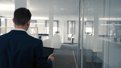 Young-caucasian-business-man-in-a-dark-blue-suit-walking-through-a-modern-office-while-looking-on-his-tablet-notes