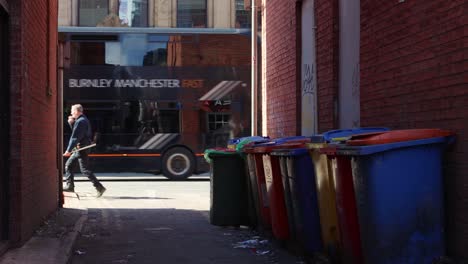 Alleyway-in-Manchester-with-colorful-bins,-passing-cars-in-background,-urban-setting