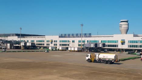 Vista-Exterior-Del-Edificio-De-La-Terminal-Del-Aeropuerto-Internacional-De-Taiwán-Taoyuan-Y-Un-Camión-Cisterna-De-Combustible-Para-Aviones-Conduciendo-Por-La-Pista-Asfaltada