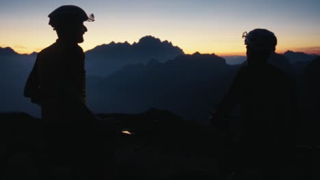 Dos-Ciclistas-Masculinos-Chocan-Los-Puños-En-La-Oscura-Puesta-De-Sol-En-Las-Montañas-En-La-Cima-De-La-Colina