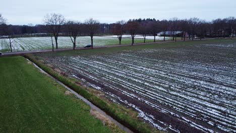 Aerial-view-of-cars-driving-down-road-in-between-agricultural-fields-covered-in-melting-snow