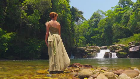 Frau-In-Einem-Kleid-Steht-An-Einem-Tropischen-Wasserfall,-Sonnenlicht-Dringt-Durch-Den-Wald,-Gelassen