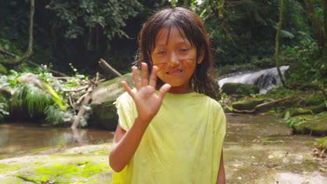 Niña-Saludando-En-La-Exuberante-Vegetación-De-Pucallpa,-Perú,-Con-Alegría-Natural-Y-Una-Cascada-Como-Telón-De-Fondo