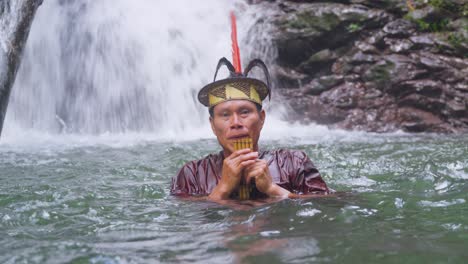 Un-Hombre-Con-Traje-Tradicional-Emerge-Del-Agua-En-Una-Cascada-En-Pucallpa,-Perú-Tocando-La-Flauta