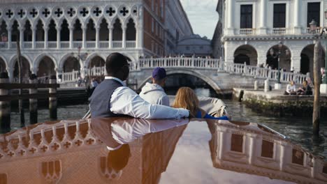 Una-Pareja-De-Raza-Mixta-Que-Realiza-Un-Viaje-Privado-En-Barco-Por-Venecia,-Italia,-Y-Está-A-Punto-De-Pasar-El-Puente-Ponte-Della-Paglia-En-El-Palacio-Ducal-Con-Turistas-En-él.