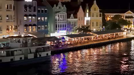 Las-Luces-Parpadean-Por-La-Noche-Detrás-Del-Ferry-Mientras-Las-Luces-Brillan-En-El-Paseo-Marítimo-De-Handelskade-Curazao-Mientras-Los-Turistas-Comen-Con-Vistas-Al-Océano.