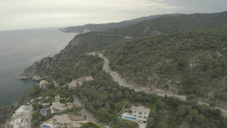 Toma-De-Drones-De-Dos-Autos-Deportivos-Conduciendo-Por-Una-Carretera-Larga-Y-Sinuosa-En-Las-Montañas-En-Un-Día-Gris-Cerca-De-Un-Lago-O-Mar-En-Italia.