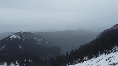 Incredible-aerial-drone-shot-of-mountains-full-of-pine-trees-and-snow,-and-a-foggy-sky-during-a-winter-snowfall