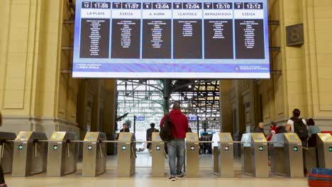 Passagiere,-Die-Durch-Drehkreuze-An-Einem-Belebten-Bahnhof-Laufen,-Fahrplantafel-Im-Hintergrund,-Tageslicht