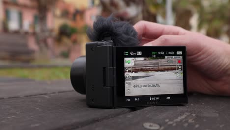 Toma-De-ángulo-Bajo-De-Una-Persona-Que-Prueba-Las-Capacidades-De-Zoom-En-Una-Pequeña-Cámara-De-Vlogging-Mientras-Descansa-Sobre-Una-Mesa-Del-Parque.