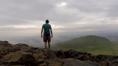 Person-arriving-at-the-summit-of-Arthur's-Seat-in-Holyrood-Park-and-overlooking-a-vast-scenic-and-mountainous-landscape-view-in-Scotland