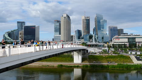 People-crossing-Baltasis-Tiltas-bridge,-Vilnius-over-Neris-river-with-Baltasis-tiltas-food-hall-in-the-background