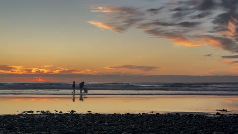 Zeitraffer-Des-Sonnenuntergangs-Mit-Silhouetten-Von-Menschen-Und-Haustieren,-Die-Den-Strand-Mit-Felsen-Im-Vordergrund-Genießen