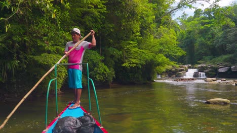 Hombre-Navegando-En-Un-Barco-Tradicional-En-Un-Río-Tranquilo-En-Oxapampa,-Perú,-Con-Exuberante-Vegetación-Y-Una-Cascada-Al-Fondo,-Durante-El-Día