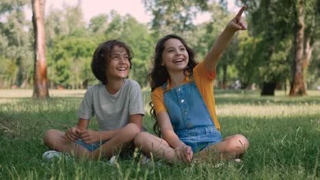 Kleines-Mädchen-Und-Junge-Reden-Und-Lachen-In-Einem-Park.-Kinder-Sitzen-Im-Gras,-Zeigen-Mit-Dem-Finger-Auf-Etwas-Und-Geben-Sich-Ein-High-Five.