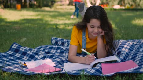 Cute-girl-in-a-park-doing-homeworks,-lying-on-the-grass-writing-in-a-notebook