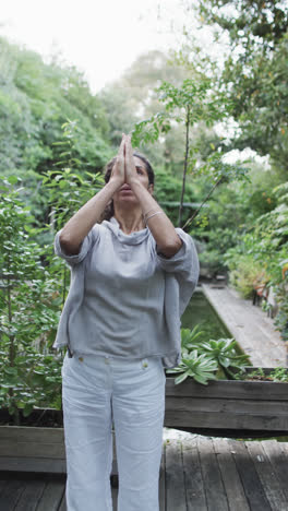 Video-Vertical-De-Una-Mujer-Birracial-Enfocada-Practicando-Yoga-En-Un-Jardín-Soleado,-Cámara-Lenta