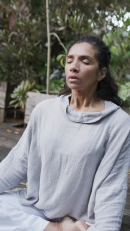 Video-Vertical-De-Una-Mujer-Birracial-Enfocada-Practicando-Yoga-En-Un-Jardín-Soleado,-Cámara-Lenta