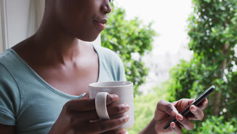 Mujer-Afroamericana-Sosteniendo-Una-Taza-De-Café-Usando-Un-Teléfono-Inteligente-En-Casa