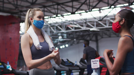 Dos-Mujeres-Caucásicas-En-Forma-Con-Mascarilla-Hablando-Entre-Sí-Y-Desinfectando-Sus-Manos-Entre-Sí