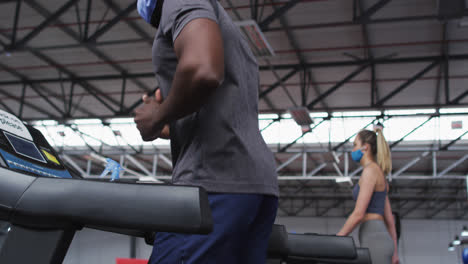 Fit-african-american-man-wearing-face-mask-and-headphones-running-on-treadmill-in-the-gym