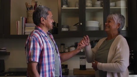 Happy-senior-mixed-race-couple-dancing-and-embracing-in-kitchen