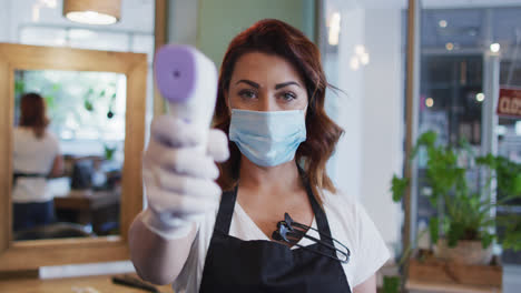 Female-hairdresser-wearing-face-mask-holding-temperature-gun-at-hair-salon