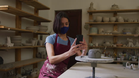 Alfarera-Caucásica-Femenina-Con-Mascarilla-Y-Delantal-Tomando-Fotografías-De-Una-Olla-En-El-Torno-De-Alfarero-En-Alfarero