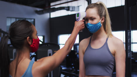 Mujer-Caucásica-En-Forma-Usando-Mascarilla-Midiendo-La-Temperatura-De-Una-Mujer-Caucásica-En-Forma-En-El-Gimnasio