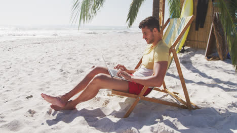 Hombre-Caucásico-Sentado-En-Una-Tumbona-Y-Usando-Su-Computadora-Portátil-En-La-Playa