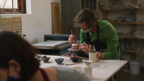 Male-caucasian-potter-wearing-face-mask-painting-on-pot-at-pottery-studio