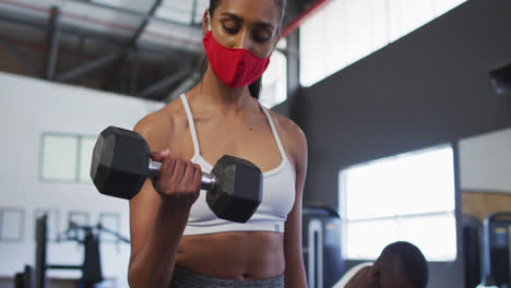 Fit-caucasian-woman-wearing-face-mask-exercising-using-dumbbell-in-the-gym