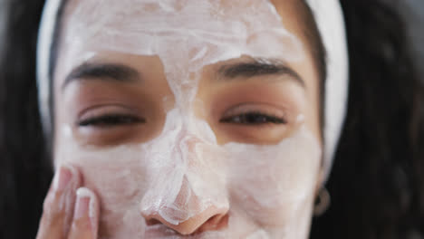 Portrait-of-smiling-mixed-race-woman-applying-face-mask-in-bathroom