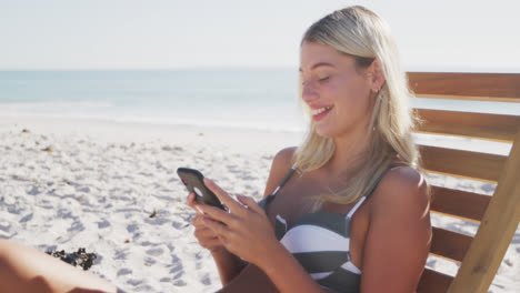 Mujer-Caucásica-Sentada-En-Una-Tumbona-Y-Usando-Su-Teléfono-Inteligente-En-La-Playa