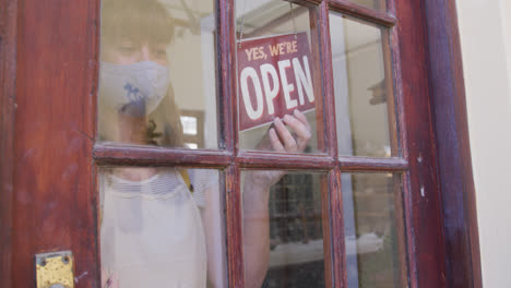 Female-caucasian-potter-wearing-face-mask-and-apron-changing-close-sign-to-open-at-entrance-of-potte