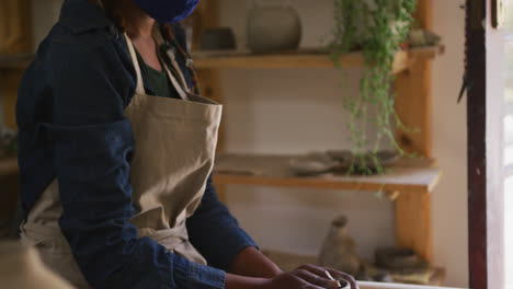 Female-potter-wearing-face-mask-and-apron-creating-pottery-on-potters-wheel-at-pottery-studio